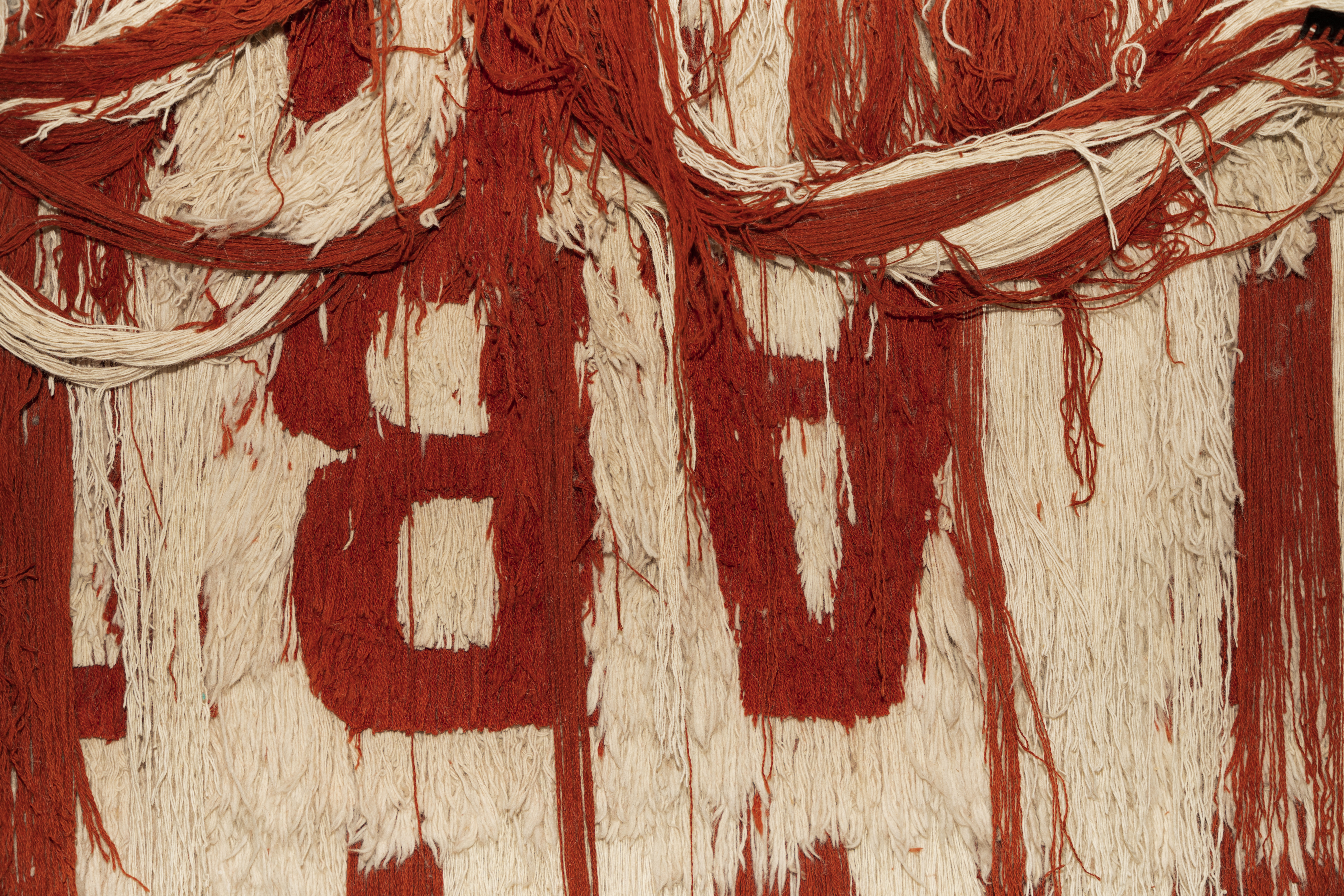 Ein Wandteppich mit zur Seite gelegten langen roten und weißen Fäden unter denen sich die auf dem Kopf stehenden Buchstaben A und B präsentieren